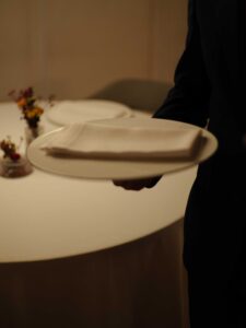 Foto 2 (Camarero con plato): Reportaje fotográfico de lifestyle en restaurante de lujo. Detalle de servicio de mesa con manos de camarero portando vajilla minimalista en un ambiente sofisticado.